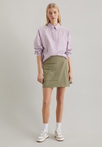 Gestreepte lavendel en witte button-up shirt gecombineerd met een olijfgroene rok met een voordezak. Het model draagt witte sokken en sneakers.