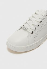 White leather sneaker with round toe, textured side panels, raised stitching details, and flat laces. Rubber sole with subtle tread pattern.