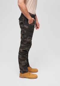 Camouflage cargo pants in dark green and black, featuring multiple pockets, reinforced knees, and a relaxed fit, paired with tan lace-up boots.