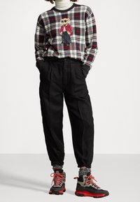 Person in schwarzen, taillierten Hosen, einem karierten Pullover mit Bärenmotiv, der über einem weißen Rollkragenpullover getragen wird, und grauen Wanderboots mit roten Schnürsenkeln.