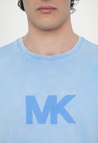 Gaiši zila kokvilnas T-kreklu ar apaļu kakla izgriezumu un lielu, izteiksmīgu "MK" grafiku nedaudz tumšākā zila krāsā. Mīksta tekstūra, brīvs piegriezums.