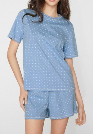 Vrouw draagt een lichtblauw polkadotshirt met korte mouwen en bijpassende short, staand met één hand op haar heup.