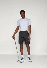 Lys grå polo med mønstrete skuldre, svarte shorts, hvit belte, hvite og marineblå sko. Står med en golfklubbe, nøytral bakgrunn.