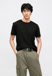 Camiseta preta de algodão com mangas curtas e decote redondo, usada com calças cargo verde-oliva e um cinto preto com ilhóses prateados.