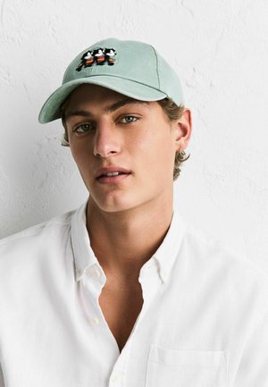 Young man wearing a light green cap with cartoon ducks and a white button-up shirt against a plain white textured wall.