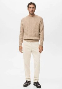 Mężczyzna w beżowym swetrze z grubym splotem, kremowych spodniach i czarnych mokasynach, stojący na tle jednolitej białej ściany.