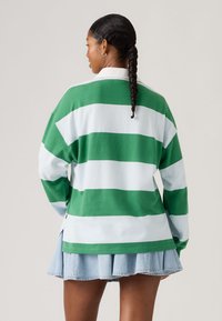 Γυναίκα με πλεξούδες, που φοράει πράσινο και λευκό ριγέ μπλουζάκι μακριάς μανίκας και ανοιχτόχρωμη φούστα τζιν με πιέτες, φαίνεται από πίσω.