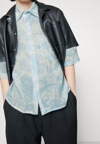 Camisa de color azul claro, con un diseño estampado y un corte relajado, llevada debajo de una chaqueta de cuero negra de manga corta. Detalles de bolsillo notables en la chaqueta.