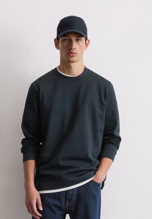 Felpa scura in misto cotone con maniche lunghe, colletto girocollo e orlo bianco. Indossata con un cappellino scuro abbinato e jeans blu.