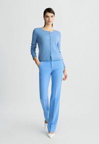 Cardigan azzurro chiaro con scollo rotondo, chiusura a bottoni e maniche lunghe, abbinato a pantaloni blu coordinati e tacchi bianchi.