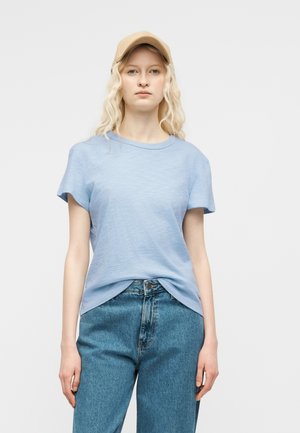 Fiatal nő hosszú szőke hajjal, bézs sapkát, világoskék pólót és kék farmert viselve, egyszínű háttér előtt áll.