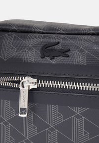 Borsa nera testurizzata con motivo geometrico, dotata di una zip argento e logo Lacoste. Materiale sintetico con una finitura liscia.