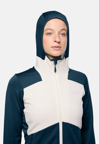 Capuchonjack in marineblauw en crème, met een textuurstof, ritsluiting en contrasterende kleurvlakken op de schouders en mouwen.