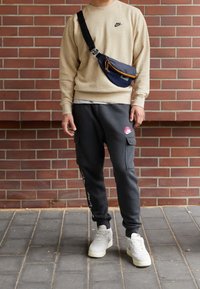 béžová mikina s logem Nike, šedé tepláky s cargo kapsami a růžovým logem Nike, tmavě modrá přes rameno taška, bílé tenisky, cihlové pozadí.