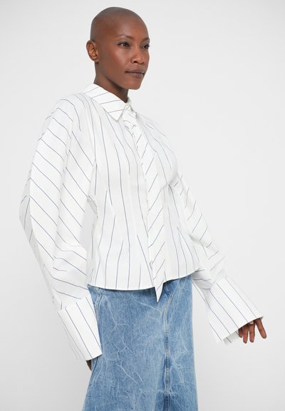 Persona calva che indossa una camicia bianca a maniche lunghe e oversize a righe con ampie maniche, abbinata a una gonna di jeans azzurri, che si trova di fronte a uno sfondo semplice.