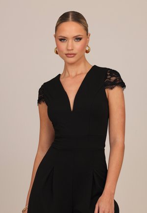 Donna con capelli pettinati all'indietro che indossa orecchini a cerchio dorati e un vestito nero con maniche corte in pizzo e una profonda scollatura a V, in piedi davanti a uno sfondo semplice.
