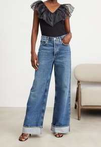 Kvinna i vida, uppvikta blå jeans, svart topp med genomskinlig volangkrage och svarta öppna klacksandaler, stående bredvid en beige stol.