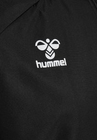 Logo bianco hummel e testo su tessuto nero strutturato, probabilmente parte di un indumento con bordo di zip visibile.