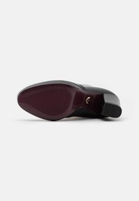 Zapato de cuero negro con tacón apilado y textura suave. La suela presenta un color burdeos con surcos y detalle de marca en dorado.