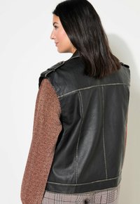 Schwarze Lederweste mit Schulterklappen, kombiniert mit einem strukturierten braunen Langarmoberteil. Die Weste hat Steppdetails und ein tailliertes Design.
