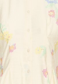 Țesătură crem cu imprimeuri florale pixelate colorate și nasturi rotunzi translucizi de-a lungul centrului.