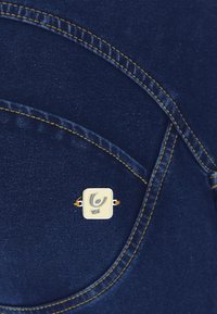 Tessuto di denim blu scuro con cuciture visibili. Presenta un distintivo quadrato con un logo stilizzato, fissato da una minuteria in tonalità oro.