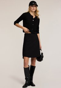 Cardigan negro corto con textura acanalada y cierre de botones, combinado con una minifalda negra y botas negras brillantes hasta la rodilla, llevando un bolso negro.