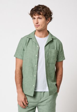 Hombre con camisa de manga corta de botonadura en verde salvia texturizado sobre camiseta blanca, pantalones a juego, de pie con una mano en el bolsillo.