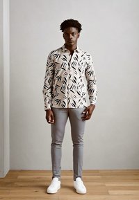 Langermet skjorte med kremfarge bakgrunn og svart blader mønster, kombinert med slim-fit grå bukser og hvite sneakers på et tregulv.