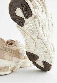 Zapatillas de deporte beige claro con superficies texturizadas, diseño voluminoso y suela de goma marrón oscuro en contraste, que presenta patrones en zigzag y ranuras.