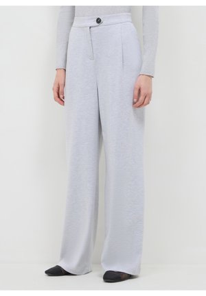 Femme portant un pantalon gris clair à jambes larges, taille haute, fermeture à boutons noirs et chaussures pointues noires sur fond uni.