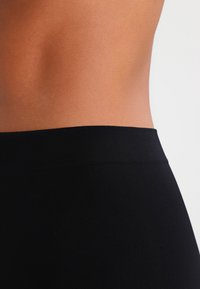 Close-up of a person’s bare midriff wearing high-waisted black fabric garment against a plain light background.