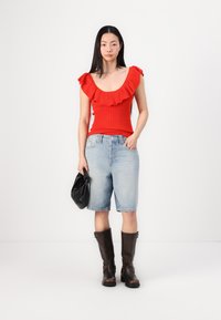 Punainen ribbed tank top, jossa on röyhelökaulus, vaaleansiniset farkkushortsit ja polven korkeudelle ulottuvat ruskeat saappaat, pitäen mustaa clutch-laukkua.