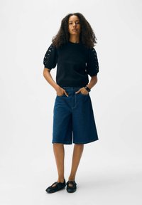 Femme aux cheveux frisés portant un haut noir à manches courtes avec des découpes, un short en denim bleu marine jusqu'aux genoux, des chaussures noires et une montre au poignet.