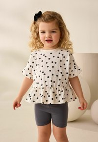 Hvid skjorte med sorte prikker, korte ærmer og peplum-hæm. Sættes sammen med grå cykelshorts. Barnet har krøllet h�år med en sort sløjfe.