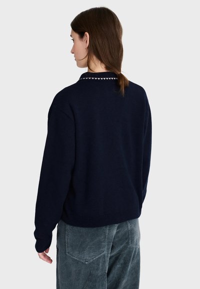 Femme aux cheveux bruns attachés portant un pull bleu marine avec un petit motif triangulaire blanc au col et un pantalon en velours côtelé gris, de dos.