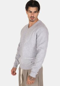 Maglioncino grigio chiaro a maglia con scollatura a V e texture a coste, caratterizzato da maniche lunghe e orlo aderente. Dettaglio del logo minimalista ben visibile in vita.