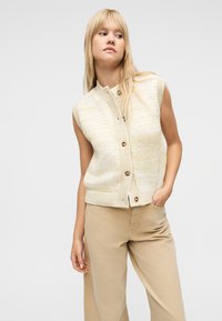 Ξανθιά γυναίκα που φοράει μπεζ πλεκτή ζακέτα χωρίς μανίκια με κουμπιά και χαλαρό μπεζ παντελόνι, στέκεται με το ένα χέρι στην τσέπη, κοιτάζοντας προς τα πλάγια.