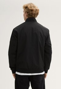 Giacca bomber nera con colletto alto, polsini e vita a coste. Realizzata in tessuto leggero, presenta una texture liscia e un design minimal.