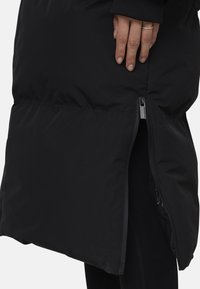 Cappotto piumino nero con una finitura opaca e liscia, dotato di una cerniera laterale e una texture leggermente imbottita, progettato per calore e comfort.