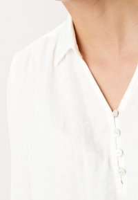 Camicia bianca in lino con colletto, patta con cinque bottoni e leggera texture; il tessuto appare liscio e leggero.