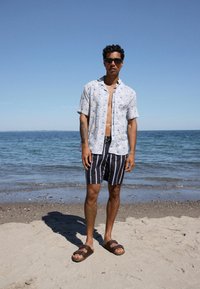 Bărbat purtând ochelari de soare, o cămașă cu mânecă scurtă cu imprimeu floral, pantaloni scurți cu dungi și sandale, stând desculț pe o plajă cu nisip lângă ocean.