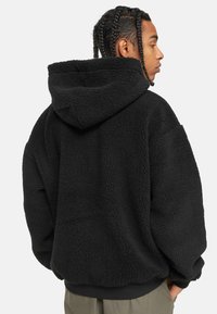 Felpa nera in pile con tessuto strutturato, design oversize e cappuccio con cordino. Polsini e orlo a coste, la vista posteriore mostra una vestibilità rilassata.
