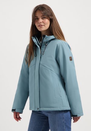 Jeune femme portant une veste d'hiver à capuche bleu clair et un jean bleu, se tenant devant un fond blanc uni.