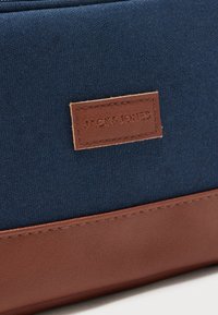 Μπλε ύφασμα και καφέ δερμάτινη λεπτομέρεια σε μια τσάντα με ένα ραμμένο καφέ δερμάτινο ταμπελάκι με ανάγλυφο το "JACK & JONES."