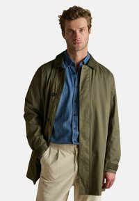 Vīrietis ar cirtainiem matiem, uzvilcis olīvu zaļu mēteļus pār zilu krekli ar pogām un krēmkrāsas korduroja biksēm, stāvot ar vienu roku kabatā.