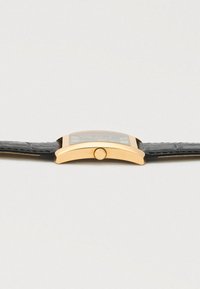 Zakřivené obdélníkové zlaté hodinky s černým koženým páskem, boční profil ukazující korunku a mírně vypouklé sklíčko.