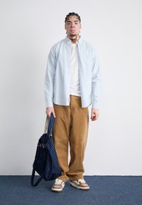 Light blue button-up shirt over a white T-shirt, paired with brown corduroy pants. Holding a blue bag with white stripes. Brown sneakers.