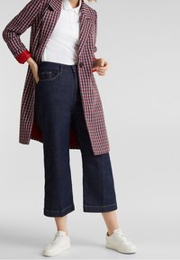 Kvinde iført en rød, hvid og marineblå houndstooth frakke over en hvid polo, mørkeblå cropped wide-leg jeans og hvide sneakers på grå baggrund.