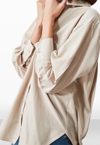 Persona con una camicia beige oversize con bottoni e maniche lunghe, jeans blu, mani vicino al viso, sfondo neutro.